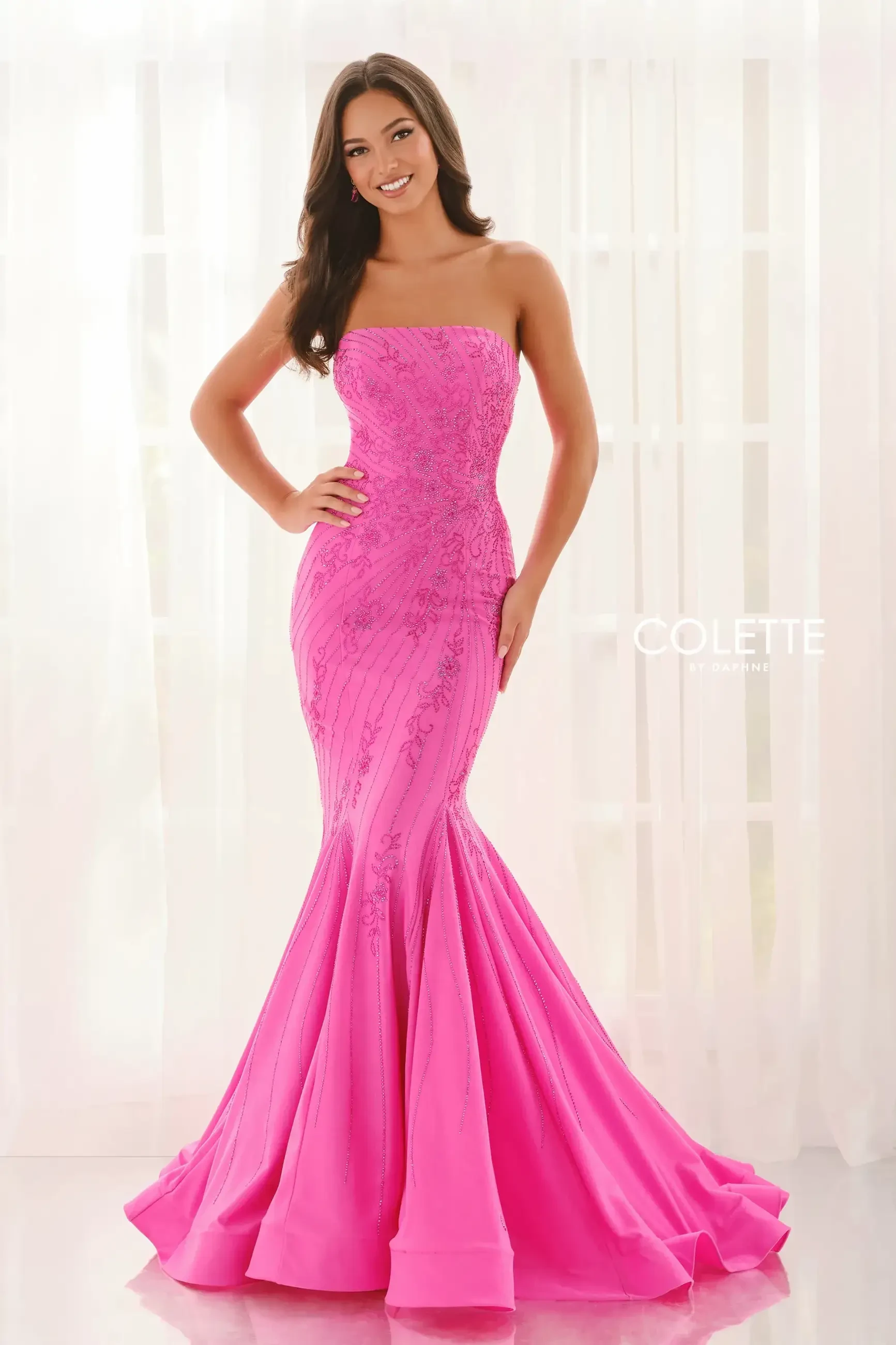 A woman is wearing a strapless pink mermaid gown with lace detailing, standing against a light backdrop.