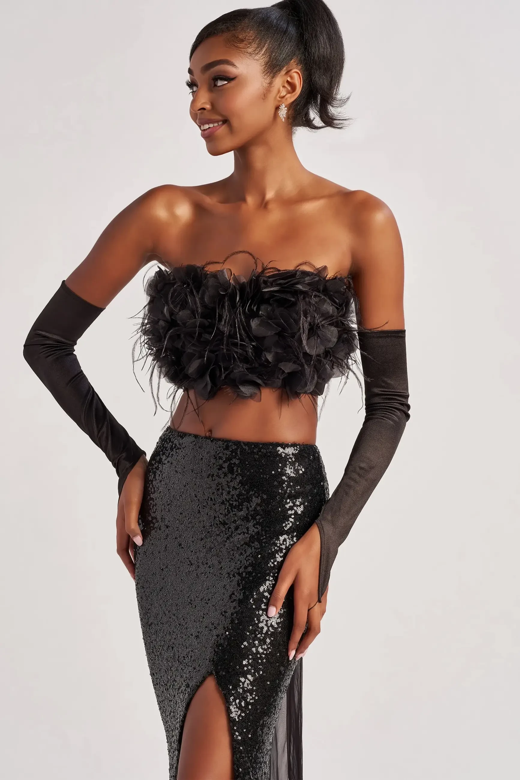 A model wearing a black strapless top adorned with feathers and a high-slit black sequin skirt, posing with a confident smile.