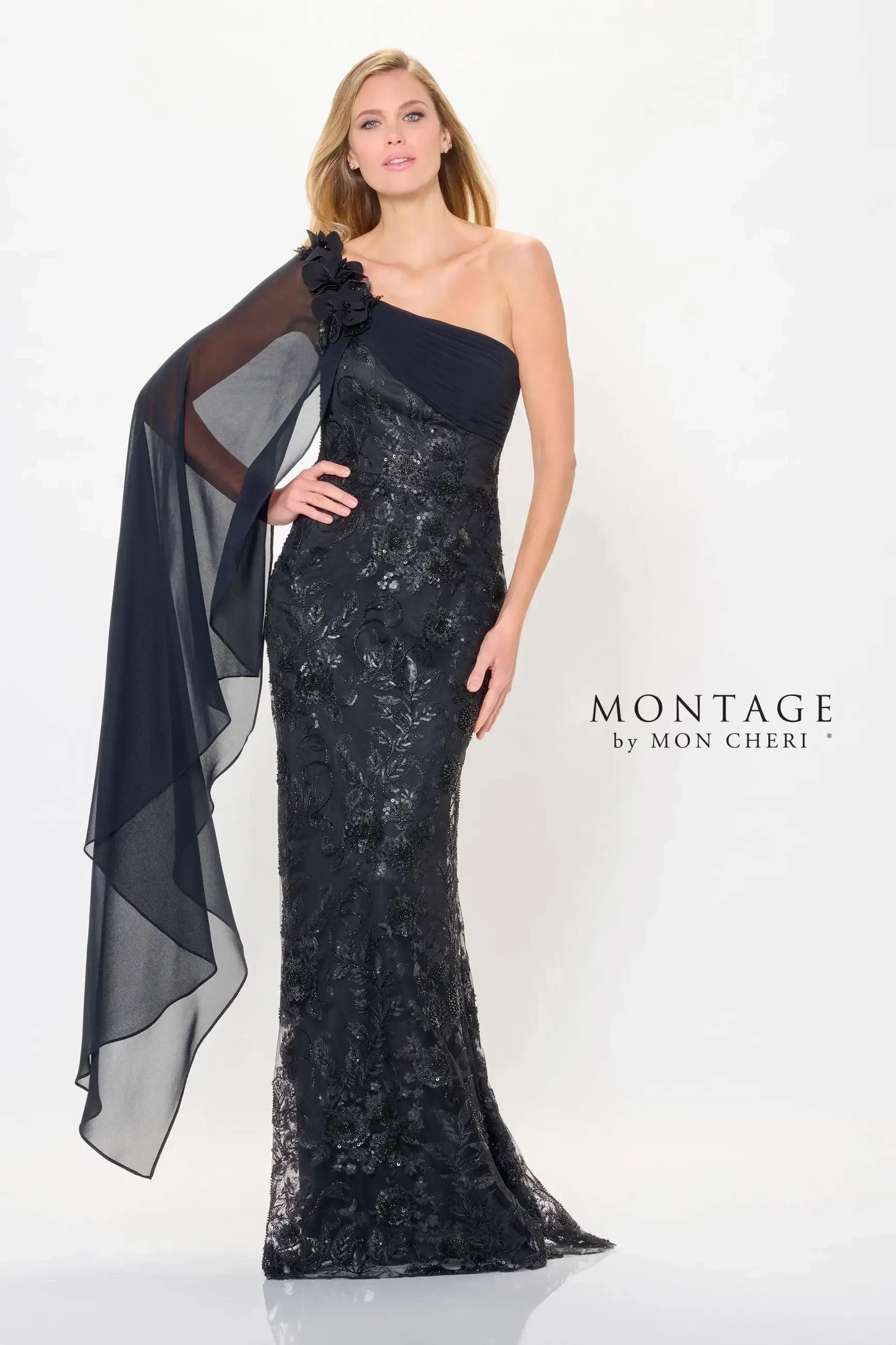 A woman poses in an elegant black gown with one shoulder and a sheer, flowing cape. The dress features intricate embellishments and a fitted silhouette.