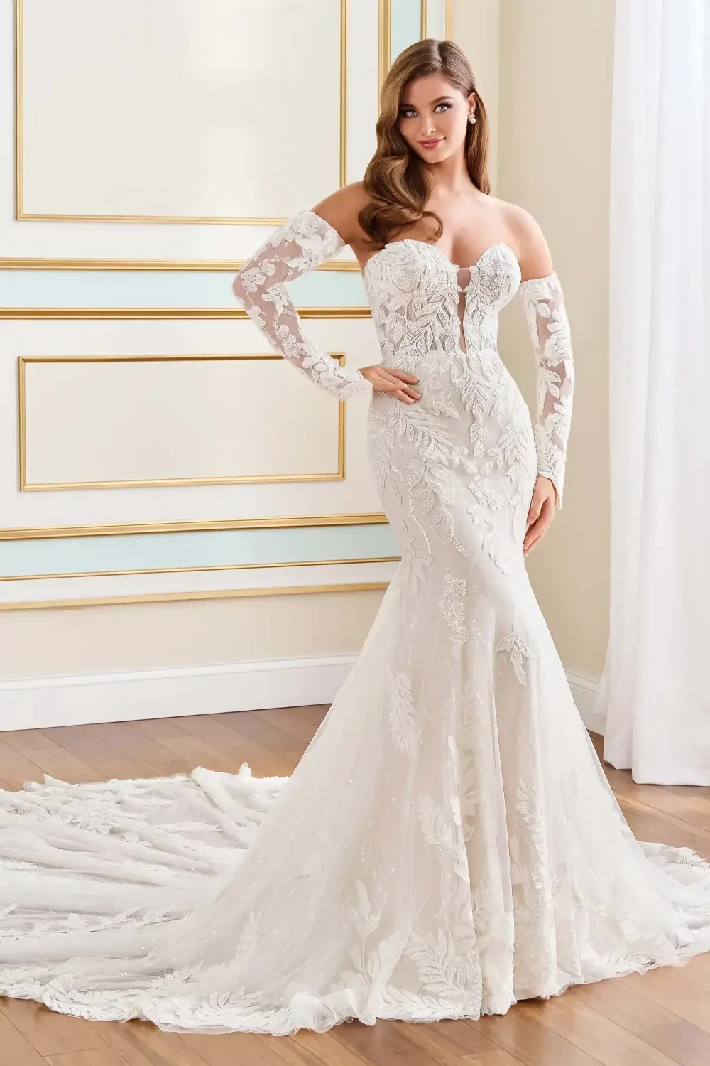 A woman poses in a formal, off-the-shoulder wedding gown with intricate lace detailing and a long train. The gown has fitted sleeves and a fitted bodice, accentuating her figure.