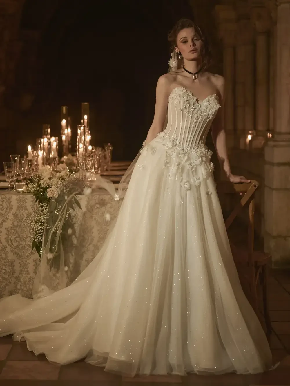 A bride in a strapless, intricately designed white wedding dress, adorned with floral details, standing near a beautifully set table with candles in a dimly lit venue.