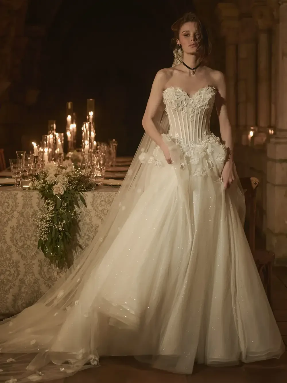 A bride in a strapless wedding dress with a corset bodice, a long flowing train, and floral accents, standing in a dimly lit room with candles and an elegantly set table in the background.